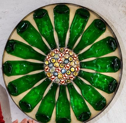 Pieza circular de muro formada por botellas de vidrio verde dispuestas en radio alrededor de un centro de tapas de colores.